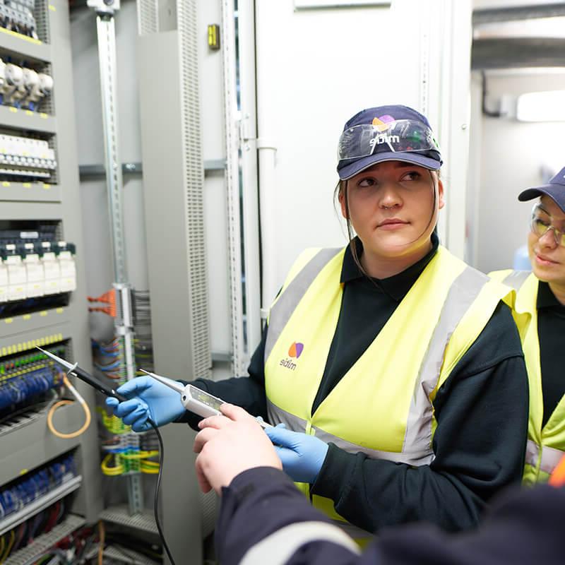 Two female engineer apprentices, wearing Mitie high vis vests and hats, 在一堵电线和保险丝墙前